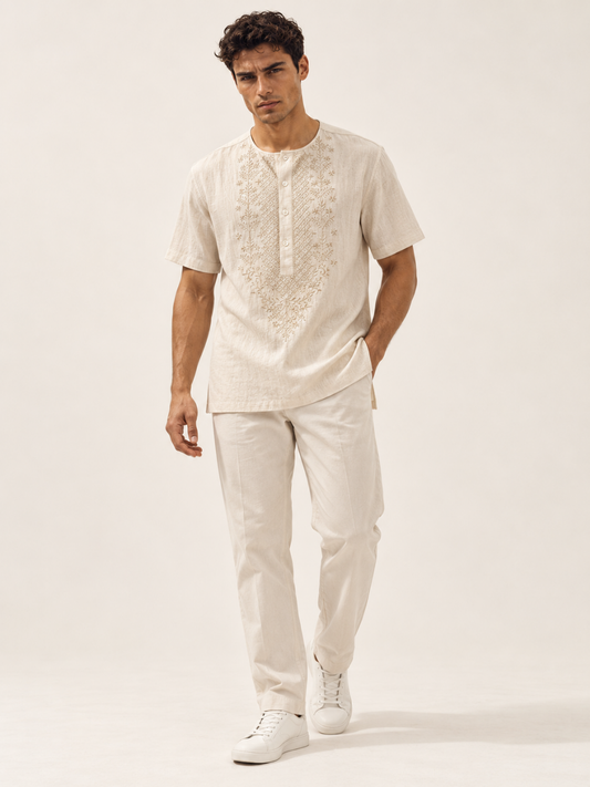 Male model wearing an ivory cream short-sleeve shirt with a round neckline, delicate beige floral embroidery and relaxed fit, styled with matching trousers and white sneakers.
