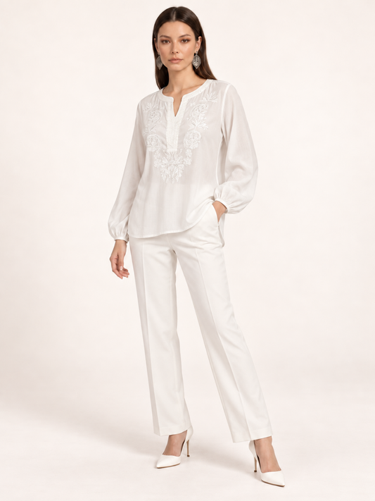 Female model wearing a white relaxed-fit blouse with floral embroidery, split round neckline and long balloon sleeves, styled with white straight-leg trousers and pointed heels.