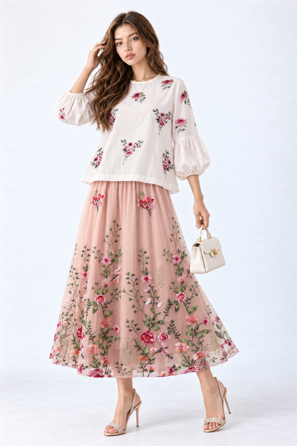 Model wearing an ivory floral embroidered blouse with round neckline and three-quarter puff sleeves, styled with a blush pink floral embroidered midi skirt, white handbag and strappy heels.