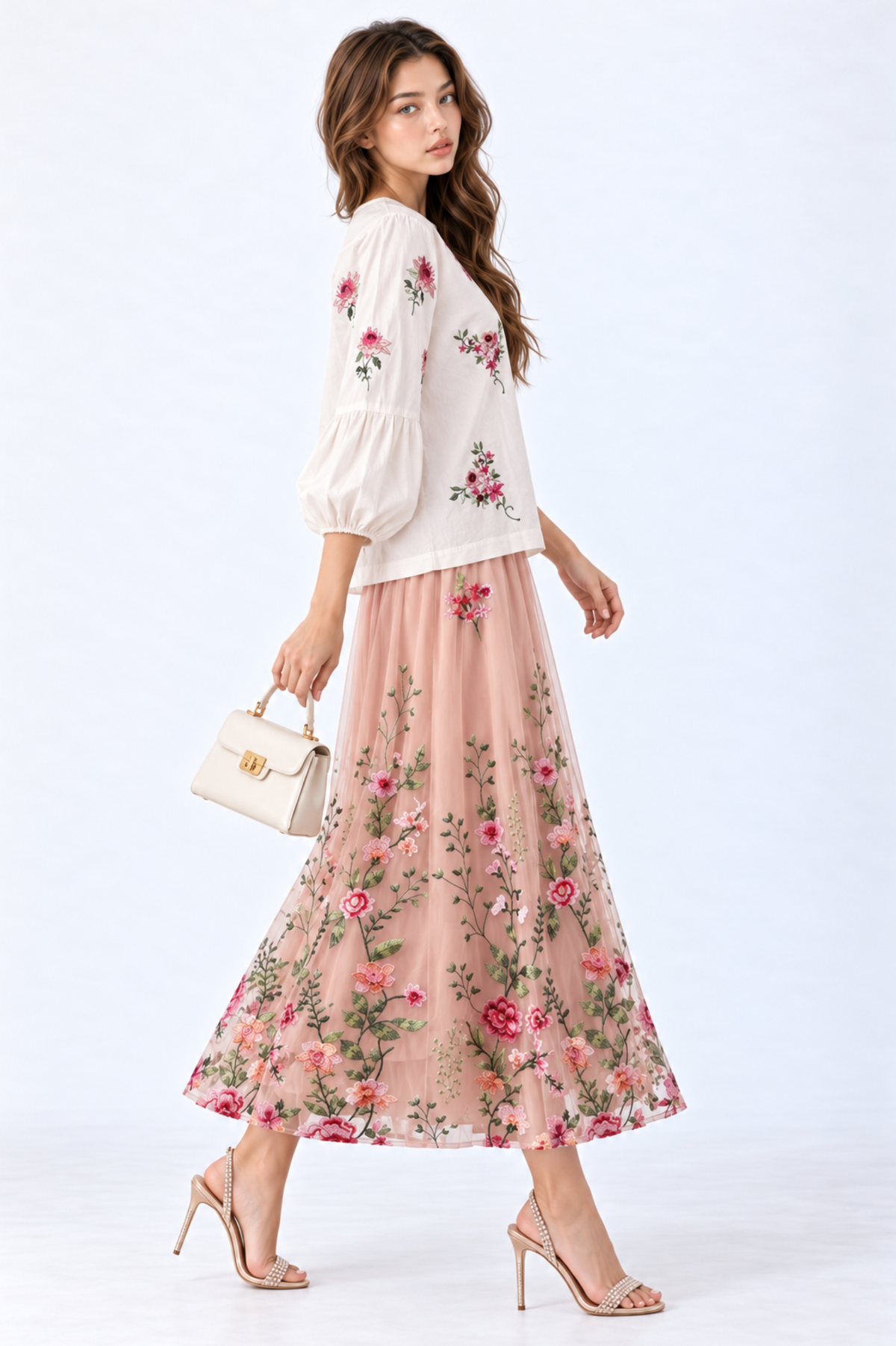 Model wearing an ivory floral embroidered blouse with round neckline and three-quarter puff sleeves, styled with a blush pink floral embroidered midi skirt, white handbag and strappy heels.