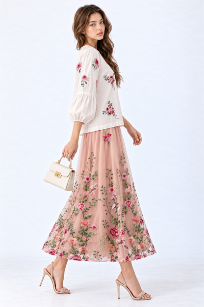 Model wearing an ivory floral embroidered blouse with round neckline and three-quarter puff sleeves, styled with a blush pink floral embroidered midi skirt, white handbag and strappy heels.