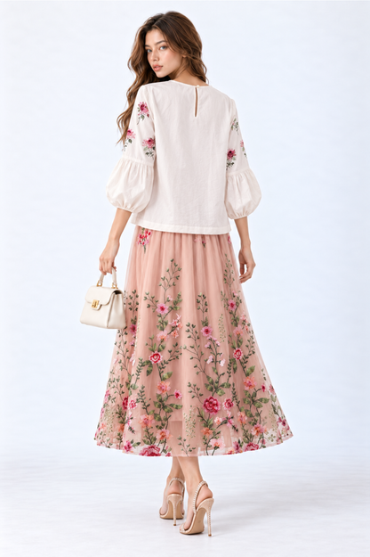 Model wearing an ivory floral embroidered blouse with round neckline and three-quarter puff sleeves, styled with a blush pink floral embroidered midi skirt, white handbag and strappy heels.