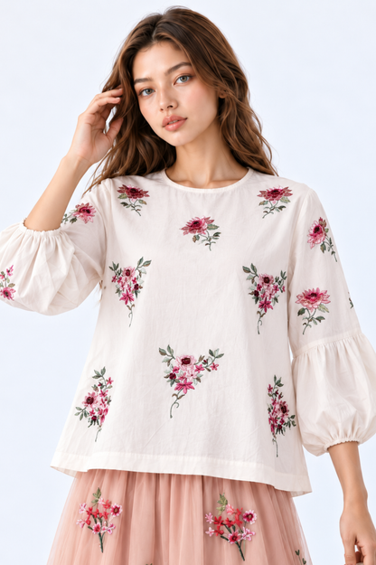 Model wearing an ivory floral embroidered blouse with round neckline and three-quarter puff sleeves, styled with a blush pink floral embroidered midi skirt, white handbag and strappy heels.