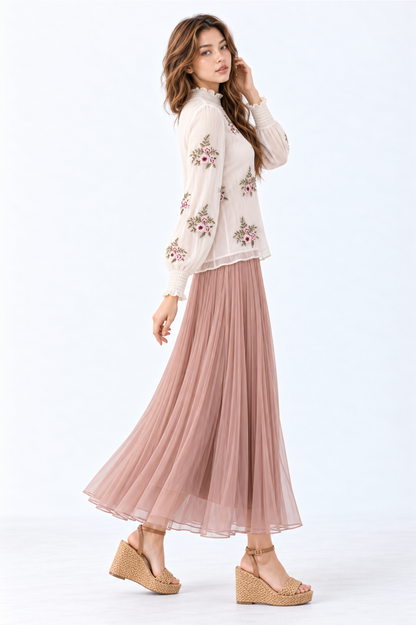 Model wearing an ivory floral embroidered blouse with high ruffled neckline, long puff sleeves and smocked cuffs, styled with a dusty pink pleated maxi skirt and wedge sandals.