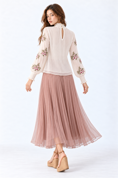 Model wearing an ivory floral embroidered blouse with high ruffled neckline, long puff sleeves and smocked cuffs, styled with a dusty pink pleated maxi skirt and wedge sandals.
