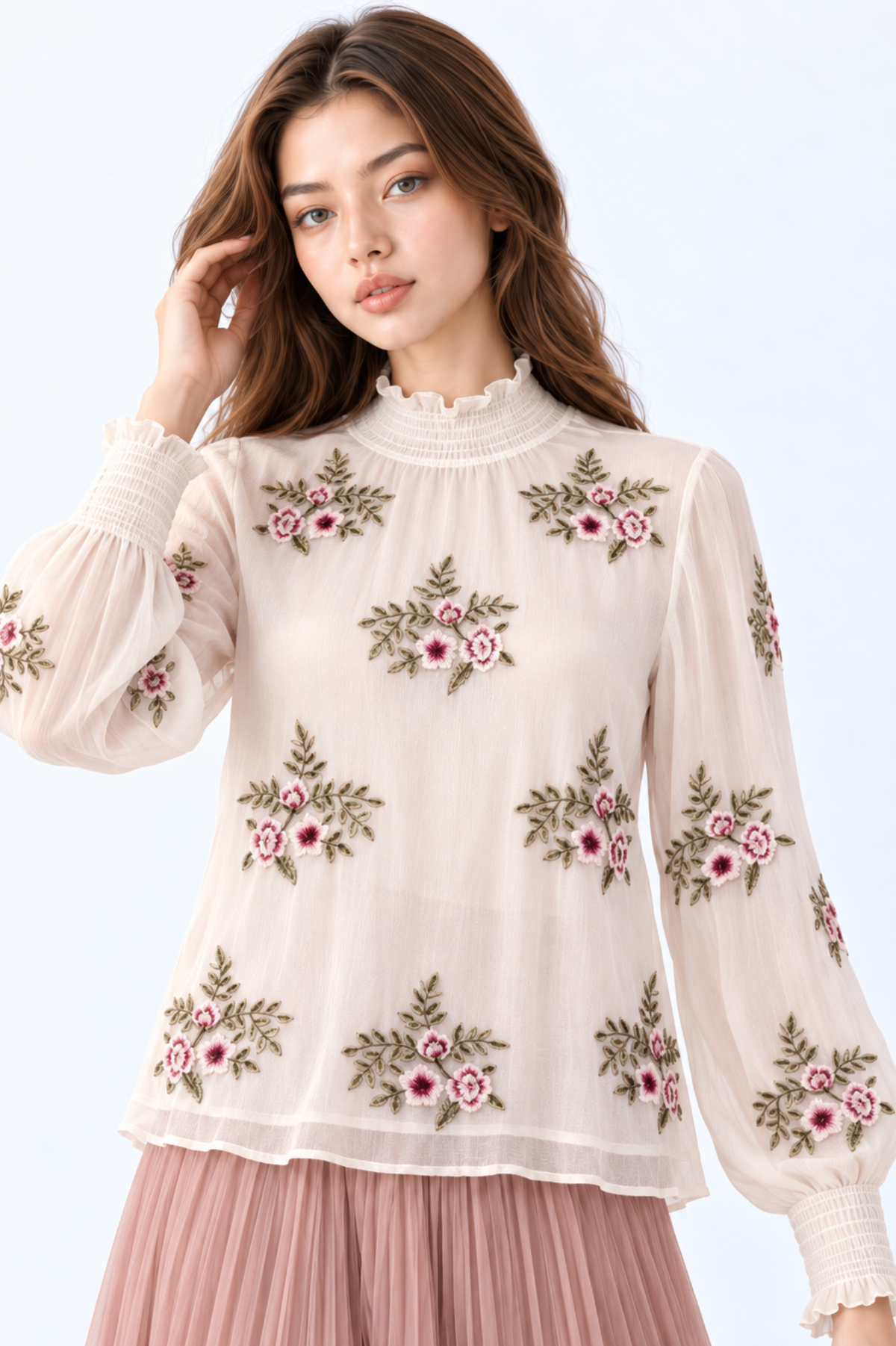 Model wearing an ivory floral embroidered blouse with high ruffled neckline, long puff sleeves and smocked cuffs, styled with a dusty pink pleated maxi skirt and wedge sandals.